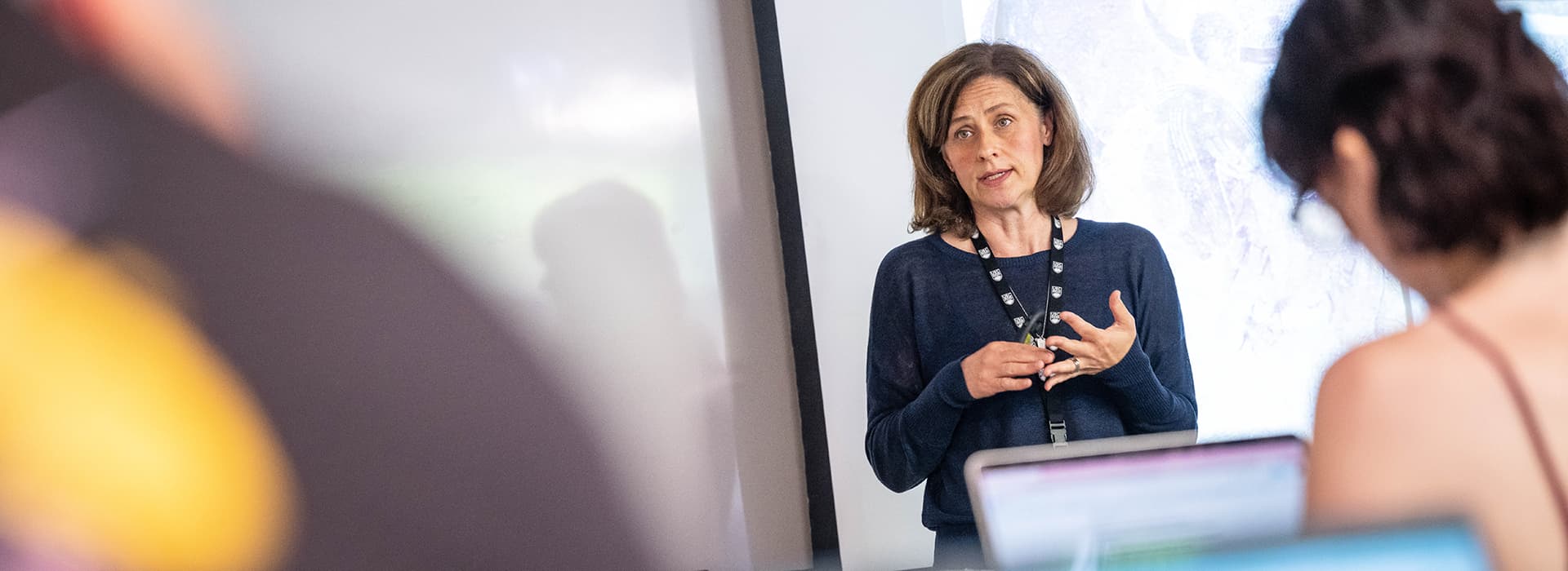 A woman stands in front of a class, with a student on their laptop in the foreground