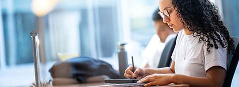 A student with curly hair and glasses in a white t-shirt, writes in a notebook