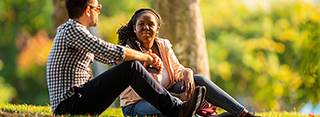 Two people in focus, sitting outside in front of two trees on a sunny day 