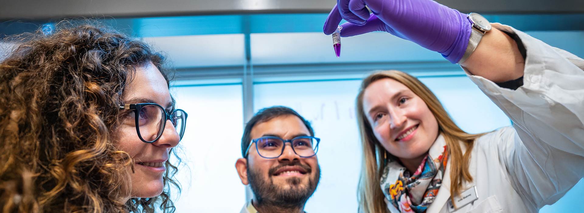Two researchers smiling, looking closely at a vile held up by a colleague with a purple lab glove 