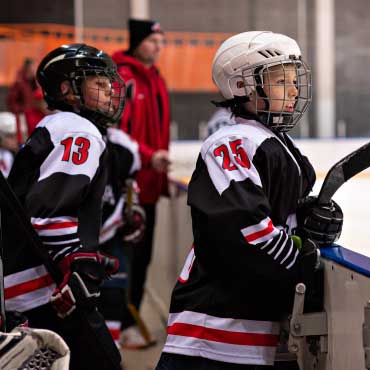 Young ice hockey players wearing helmets and uniforms hold hockey sticks and look towards the ice from the side of a rink.